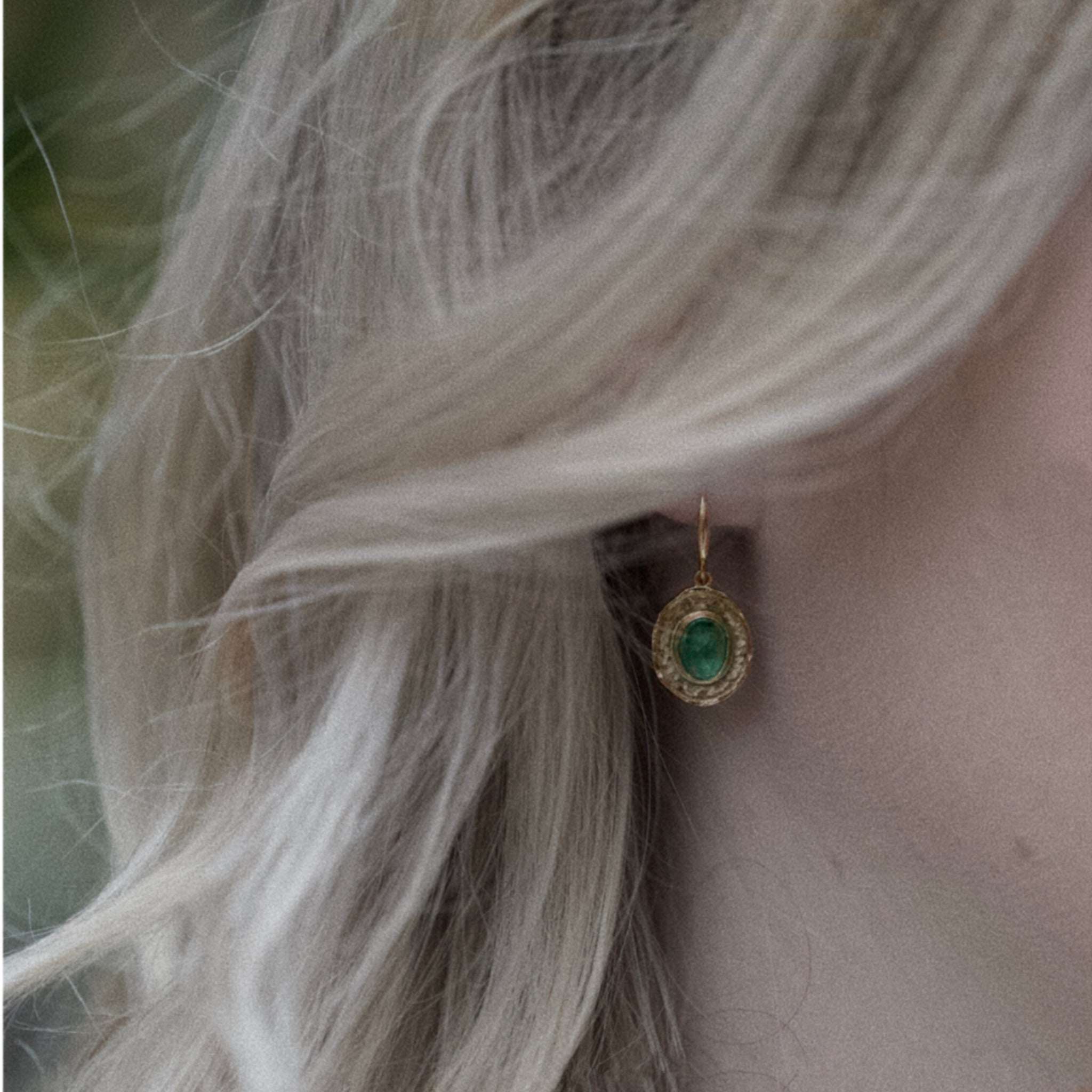 Close-up of a person wearing Florin Emerald Earrings featuring hammered gold and a vibrant blue-green emerald gemstone.