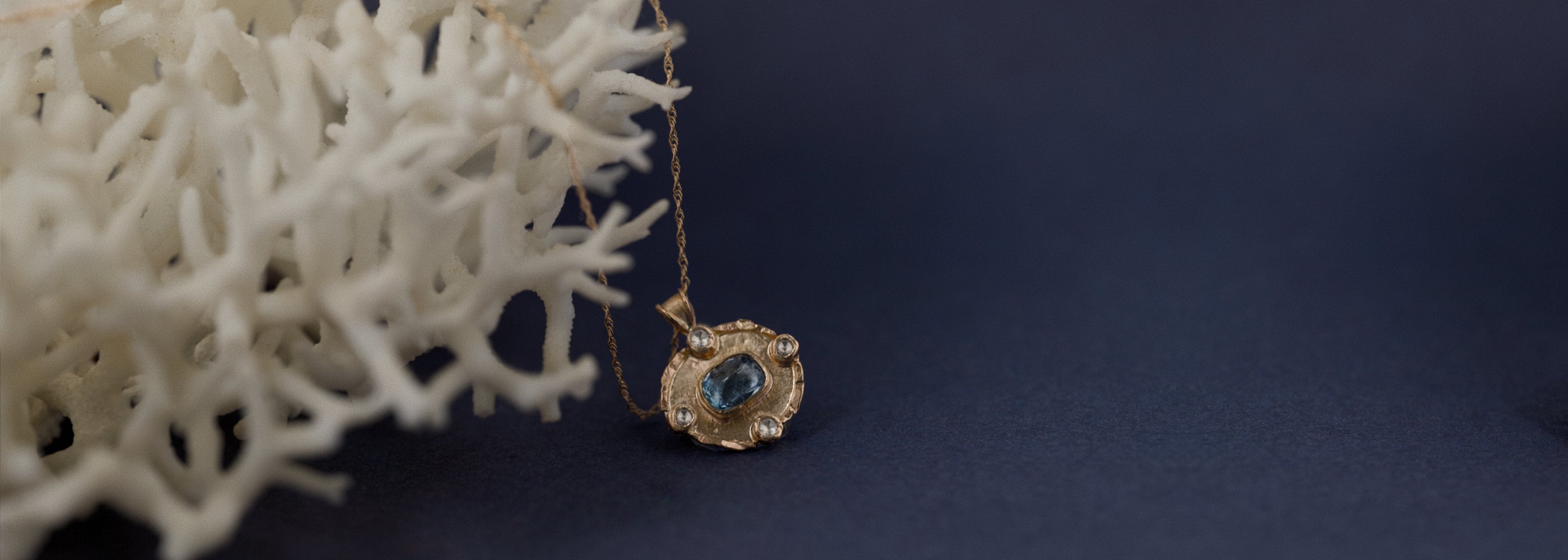 Aquamarine pendant in textured gold beside white coral on deep blue background.